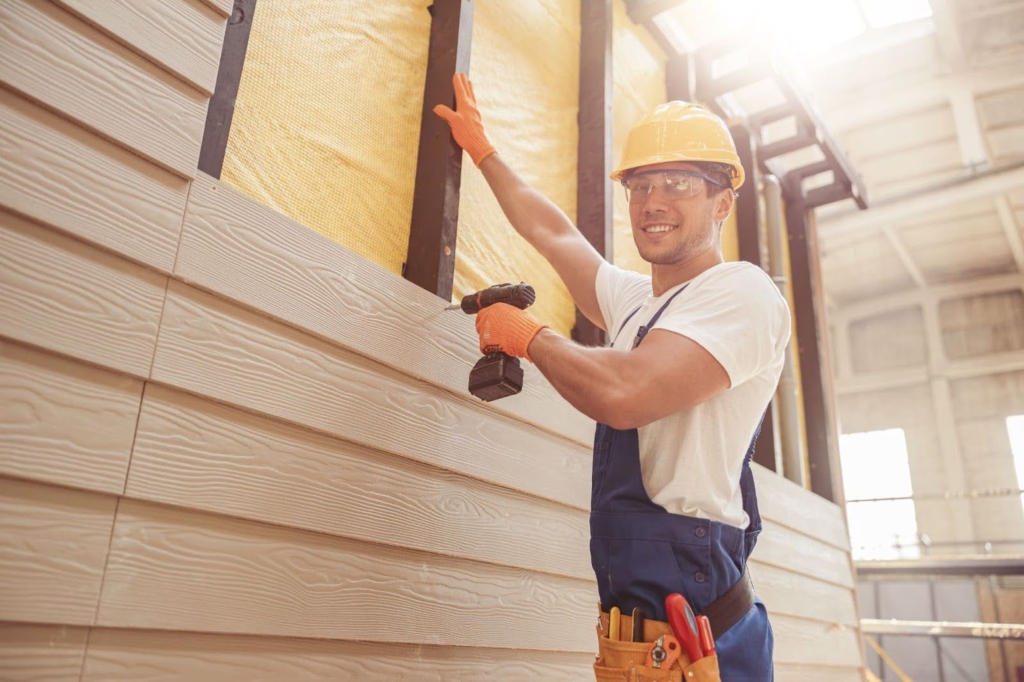 A man in a hard hat installs siding with a drill.