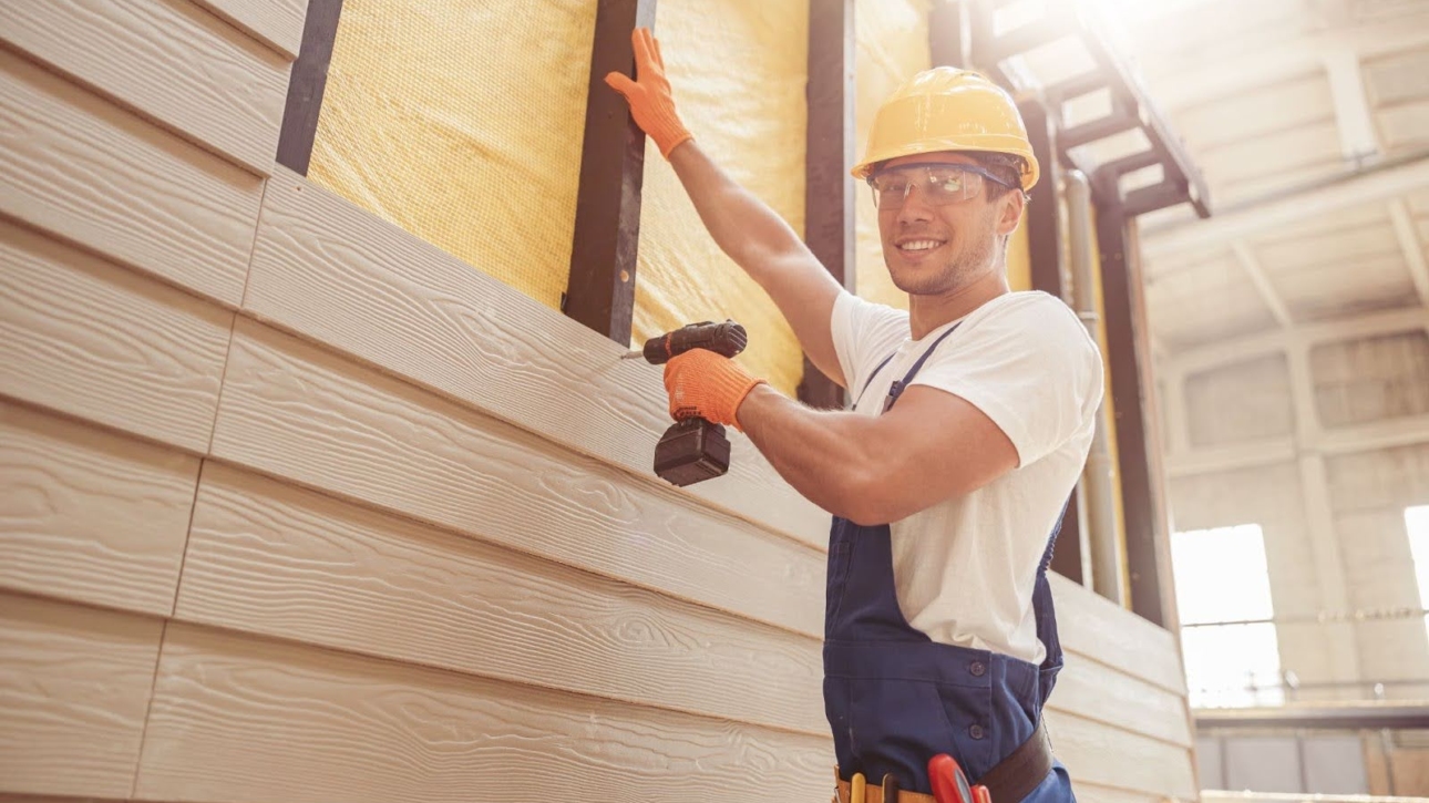 A man in a hard hat installs siding with a drill.