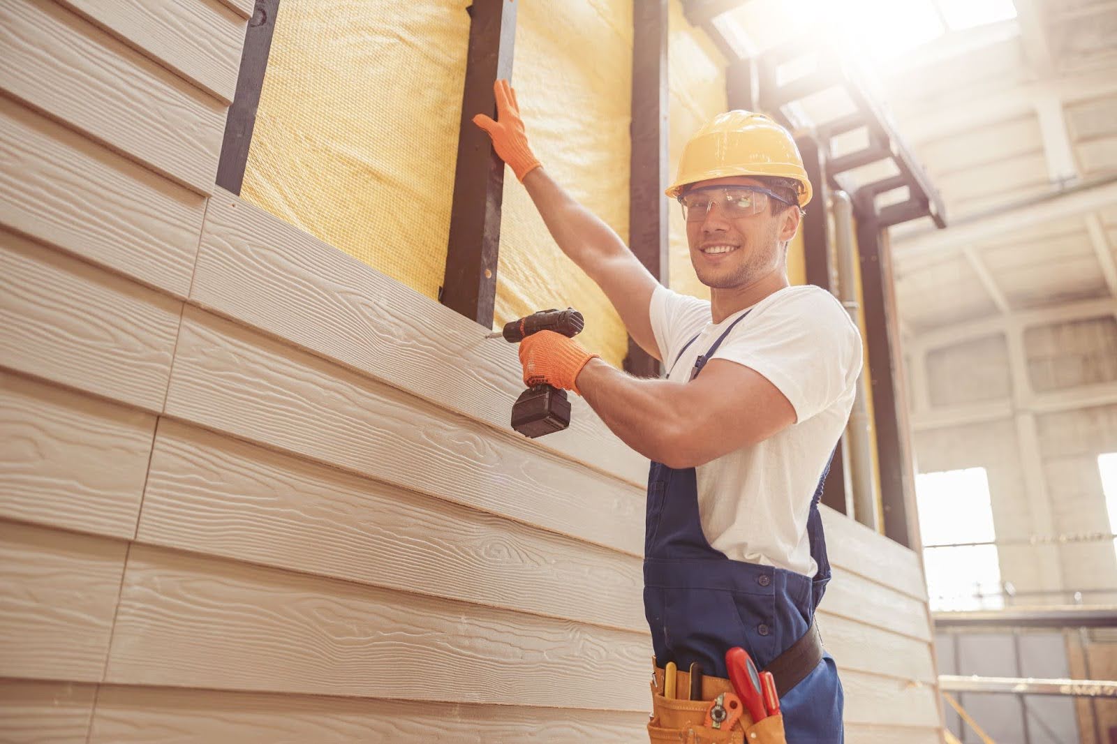 A man in a hard hat installs siding with a drill.